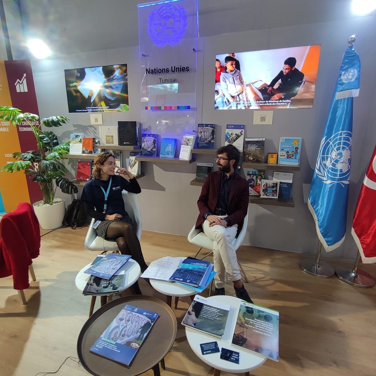 Dr. Meriem Rebbani engaged in a discussion at a United Nations event, seated with a colleague in a professional setting, with UN flags and informational materials visible.